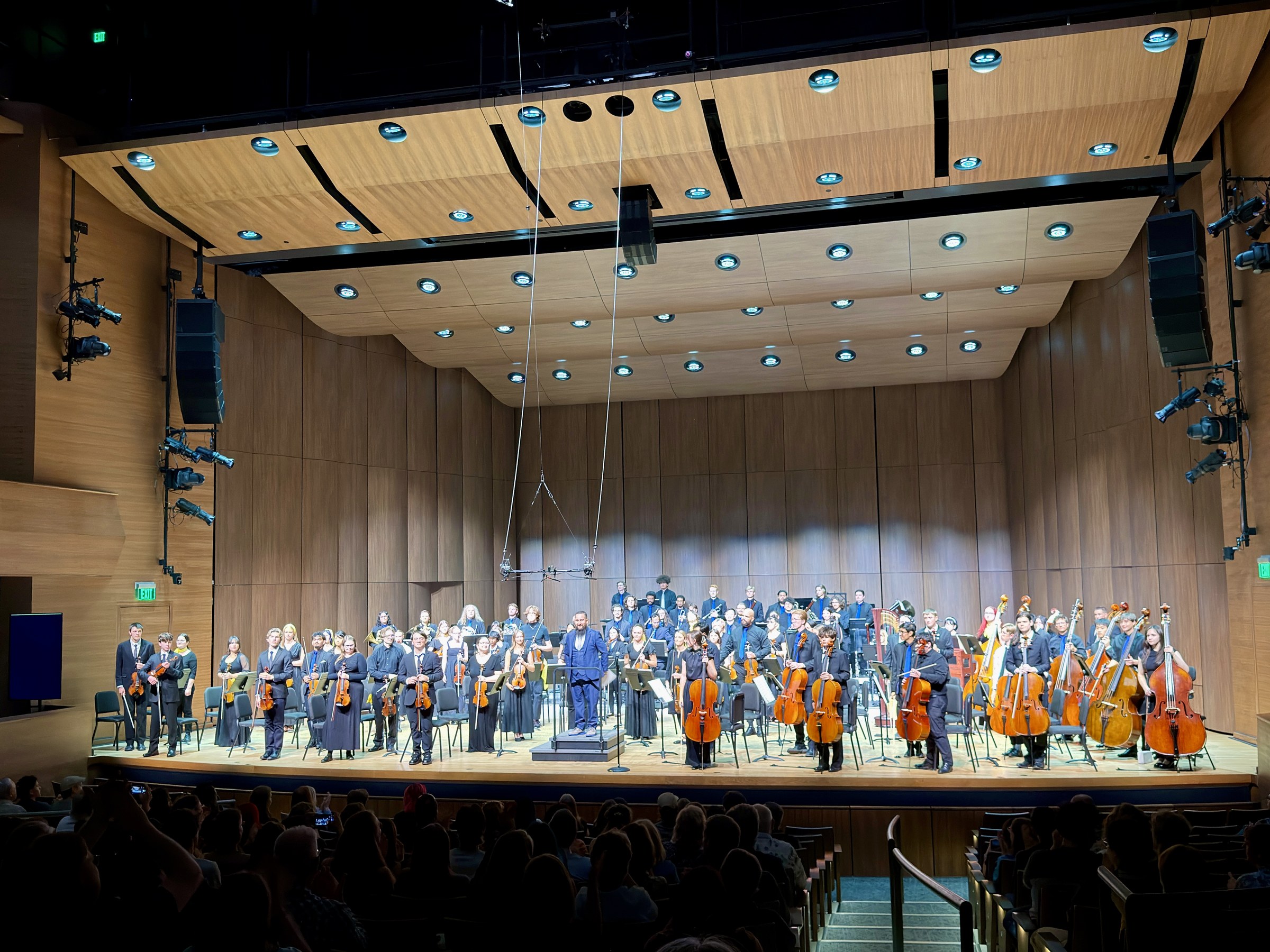 NoCoYO’s full orchestra standing together on a concert stage, with strings in front and winds, brass, and percussion behind them